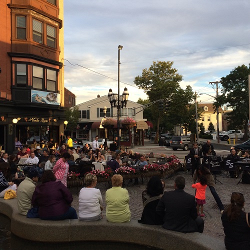 Photograph of more than 40 individuals sitting in a public park and an adjacent outdoor restaurant seating area listening to musicians play.